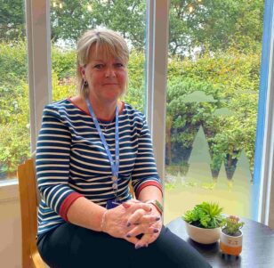 A light haired woman sits on a chair facing the camera, her hands are clasped together resting on her knee