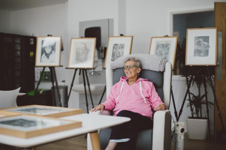 Colleen wearing a pink top, sitting in a chair in front of her displayed artwork