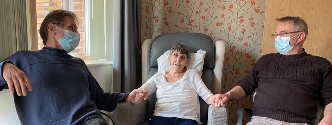 Ann sat in chair at the hospice with sons sat either side of her holding hands