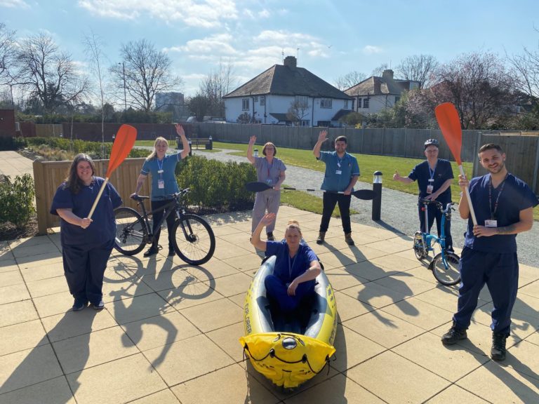 Seven members of care staff wearing blue scrubs are outside holding an oar, sitting on a bike or within a kayak looking to camera and waving
