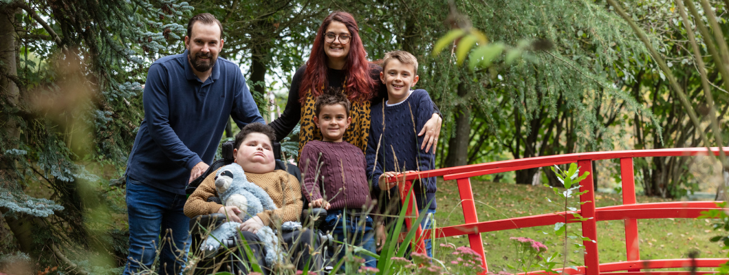 Teenage boy in a wheelchair holding sloth toy, with parents behind him and brothers stood next to him. Family photo in the garden at the hospice