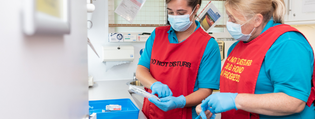 Two nurses wearing blue tops with red drug round vests on preparing medication