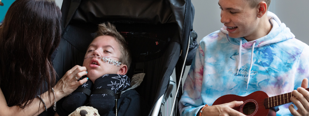 boy in wheelchair at the hospice with mum next to him and brother playing ukulele