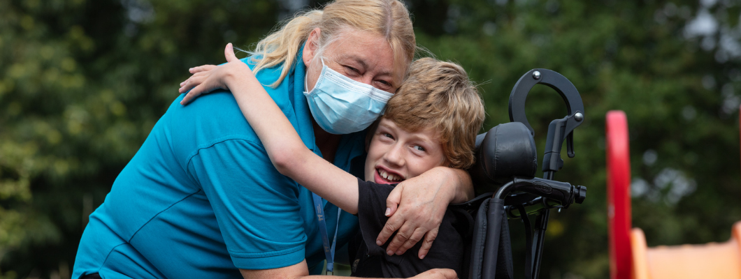 Boy sat in wheelchair hugging nurse outside at the hospice wearing mask