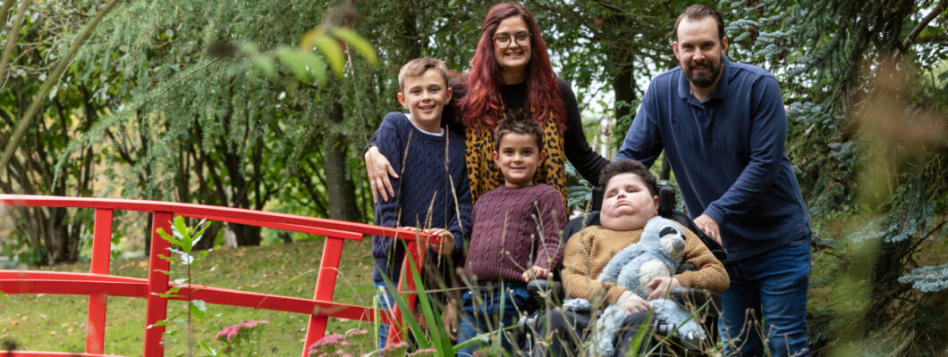 boy holding sloth toy sat in wheelchair in garden with family around him at Little Havens