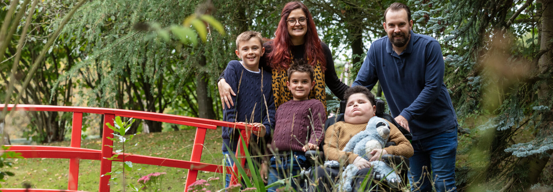 boy holding sloth toy sat in wheelchair in garden with family around him at Little Havens