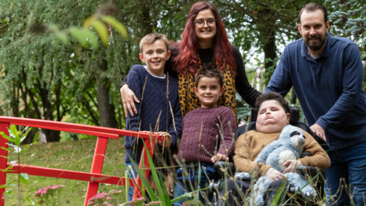 boy holding sloth toy sat in wheelchair in garden with family around him at Little Havens