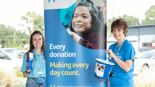 two people wearing havens blue tops by tall banner holding bucket collections