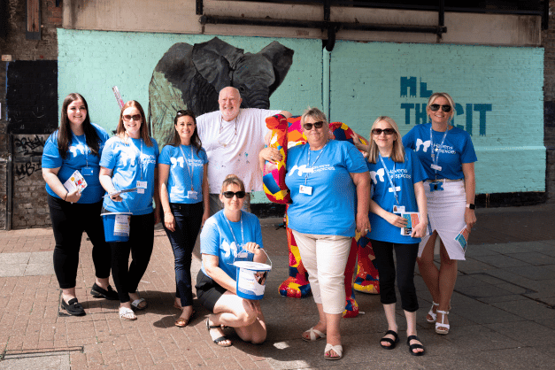 A group of people wearing blue havens hospices t shirts smiling to camera. A man wearing a white t shirt stands in the middle. In the background a mural of elephant
