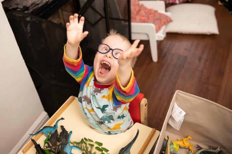 A little boy called Noah wearing glasses and wearing a dinasour top is laughing lifting his hands high in the air