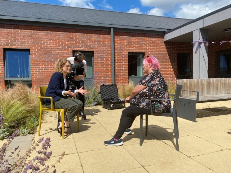 A female sits on a chair, with pink hair and band. Looks towards a TV camera with another female in a chair and a male camera operator