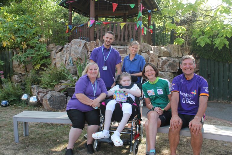 Three care team members wearing purple t shirts sit and stand around a young girl with glasses and white legging,s using a wheelchair. Francesca and Tony, the cyclists, are wearing cycling gear and sitting next to the young girl. All are looking to camera. In the background are gardens.