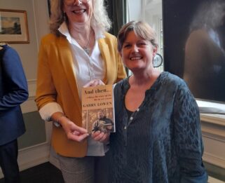 Two women standing, holding a book and smiling to camera. One wearing a yellow jacket and blond hair, the other wearing a dark blue top with short brown hair