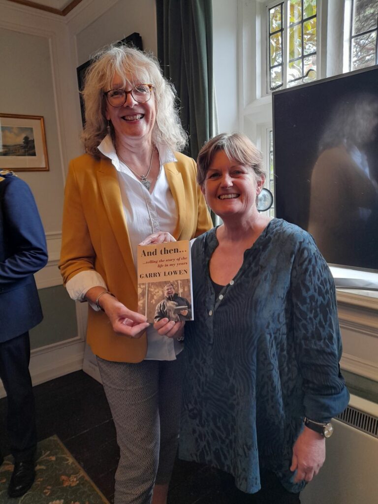 Two women standing, holding a book and smiling to camera. One wearing a yellow jacket and blond hair, the other wearing a dark blue top with short brown hair