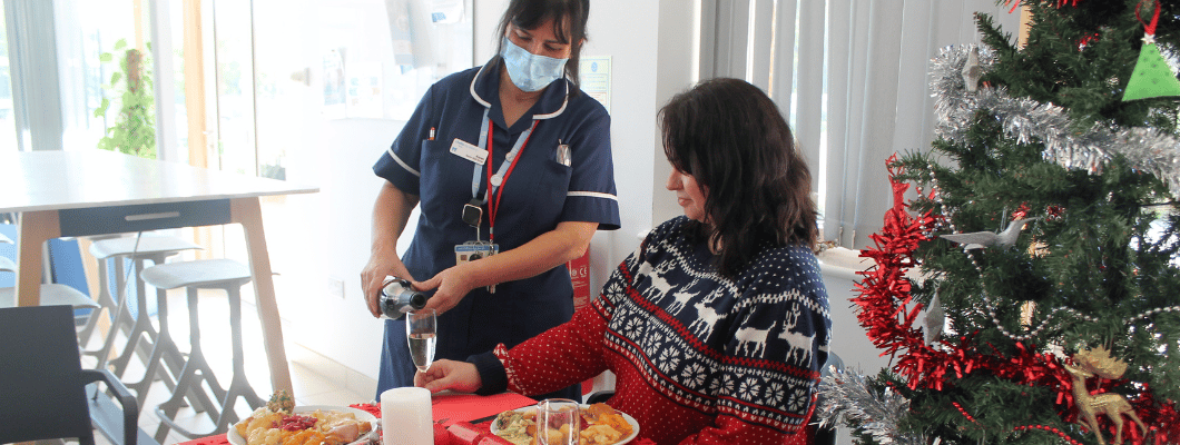 Lady sat at table wearing christmas jumper with nurse wearing a mask pouring her a drink