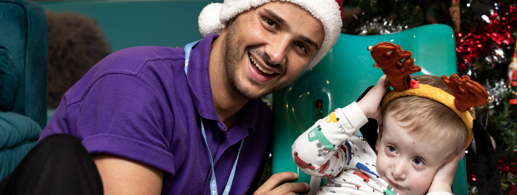 Little havens patient sat in green chair with christmas headband and male carer next to him wearing a santa hat out of shot