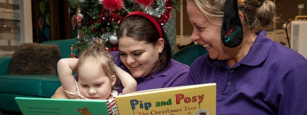 Two members of Care Stafff and a Little Havens child are dressed in festive head gear and reading a book together. Christmas decorations can be seen in the background.