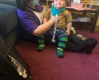 Elijah sitting on Amanda's lap. Looking up and smiling. Amanda is wearing a purple top and sitting on the floor with her back against the sofa. She is wearing a face covering.