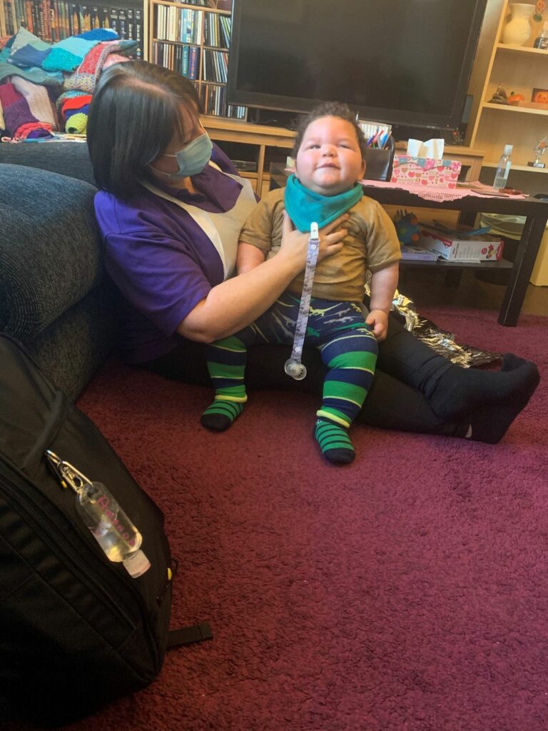 Elijah sitting on Amanda's lap. Looking up and smiling. Amanda is wearing a purple top and sitting on the floor with her back against the sofa. She is wearing a face covering.