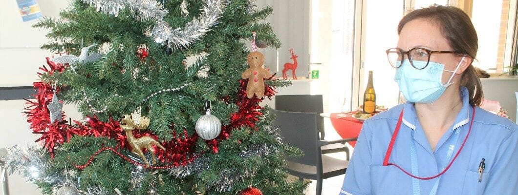 A Fair Havens nurse in uniform, wearing a PPE mask, sitting in front of a Christmas tree