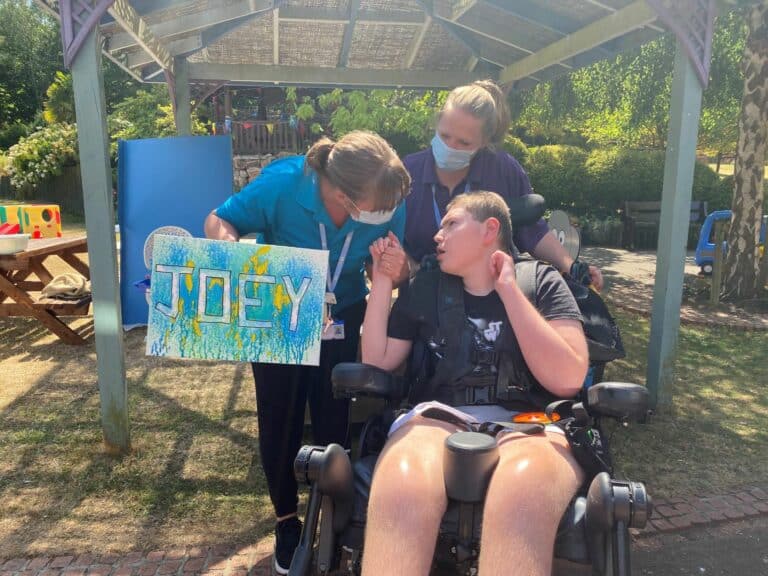 Joey looking towards a care team member whilst another care team member is behind his wheelchair. They are holding a piece of artwork which says 'Joey'