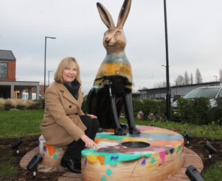Sandra Karslake wearing a log beige coat kneels down by the har sculpture and points to a plaque featuring Paul's name
