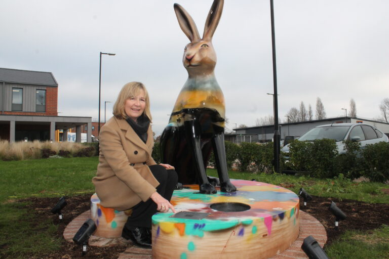 Sandra Karslake wearing a log beige coat kneels down by the har sculpture and points to a plaque featuring Paul's name