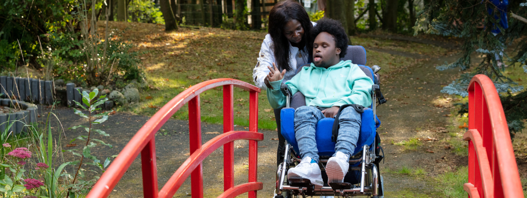 William, a teenage boy, wearing mint green hoody being pushed in his wheelchair over a red bridge in the garden