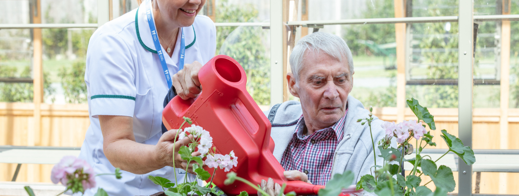 Older man sat in the garden watering plants with therapist in white uniform supporting the watering can