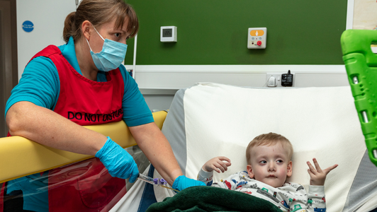 young boy in bed watching his ipad while nurse wearing a red vest is delivering medication via a tube