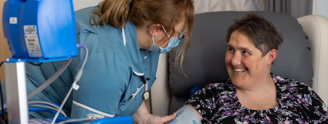 Amanda, patient at Fair Havens, sat in a chair wearing a blood pressure cuff. Nursing Assistant wearing glasses and mask stood next to her with her hand gently on Amanda's arm