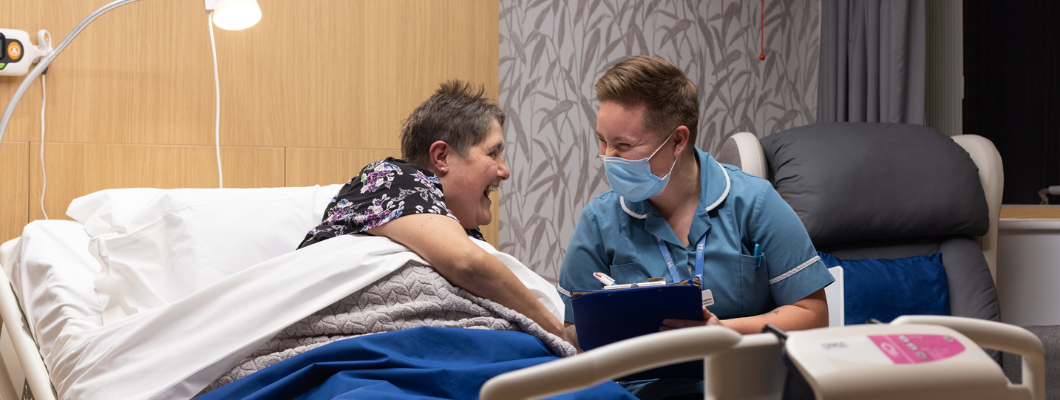 Fair Havens patient in bed in room with nursing assistant sat next to her making notes, both looking at each other and laughing