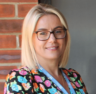 A lady with blonde hair wearing glasses stood outside by a brick wall smiling at camera