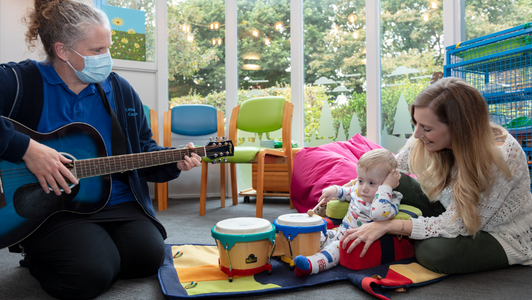 young boy sat on the floor with mum playing drums, music therapist on the side playing guitar