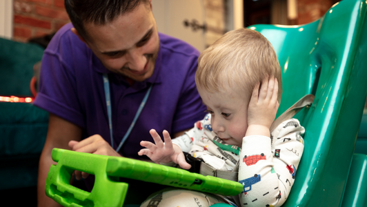 Little Havens child sat in a green seat holding his Ipad with his hand leaning on his head. Care Team member wearing blue top next to him
