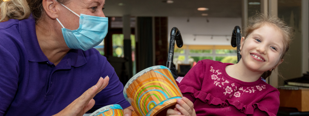 Child leaning to the side mouth opening laughing with Care Team member sat next to her wearing a mask banging on a drum