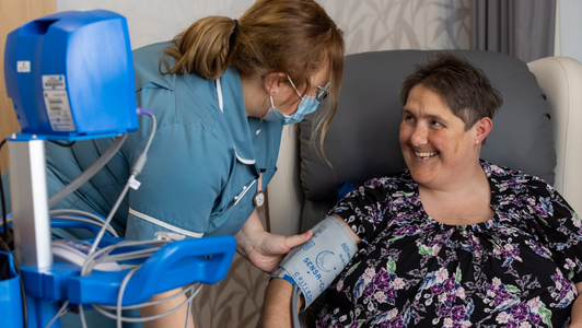 Amanda, patient at Fair Havens, sat in a chair wearing a blood pressure cuff. Nursing Assistant wearing glasses and mask stood next to her with her hand gently on Amanda's arm