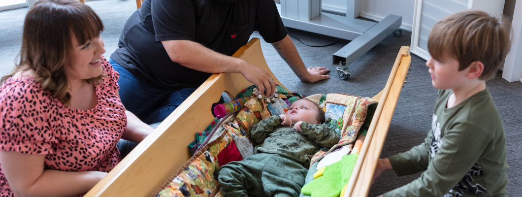 Young baby laying in wooden sound cradle on floor with musical instruments next to him. Older brother and mum sat either side smiling at the child