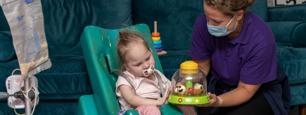 Young girl with dummy sat in green supportive chair on the floor, care team member is next to her wearing a mask and holding out a merry-go-round toy. In the corner of the shot is a milk pump feed set up