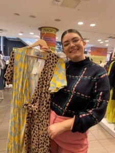 Amy Lorenz wearing glasses smiling at the camera holding up a dress on a hanger