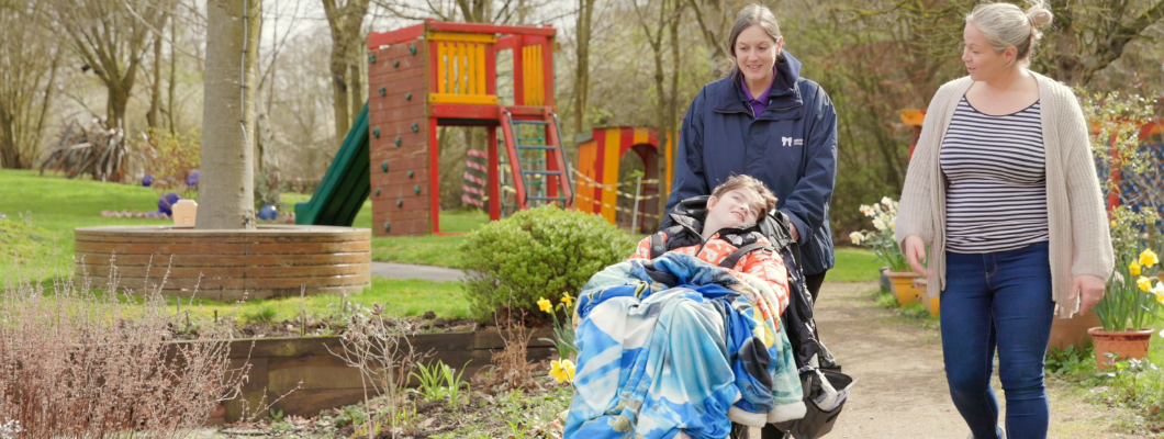 A boy in a wheelchair being pushed around the garden with a carer with his mum stood next to him smiling at him