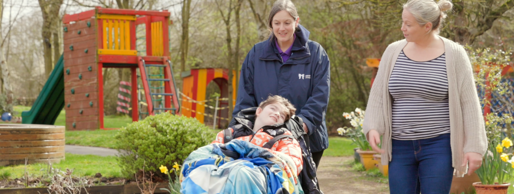 A boy in a wheelchair being pushed around the garden with a carer with his mum stood next to him smiling at him