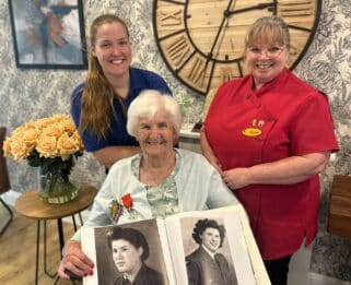 Carys and Sarah standing behind Joan who is sitting down with images of his in the Woman's Auxiliary Air Force