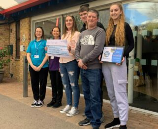 a famiily standing outside Little Havens reception with two members of the hospice's care team holding a cheque and a framed certificate