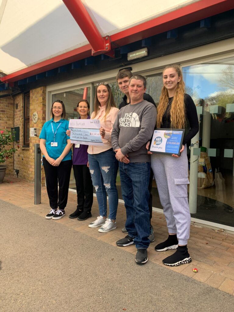 a famiily standing outside Little Havens reception with two members of the hospice's care team holding a cheque and a framed certificate