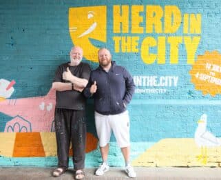Male artists John Bulley and Karl Sims standing against a colourful background featured Herd in The City logo and pink elephant holding an ice cream