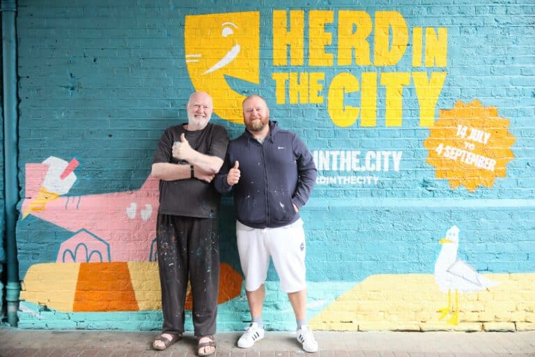 Male artists John Bulley and Karl Sims standing against a colourful background featured Herd in The City logo and pink elephant holding an ice cream