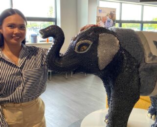 Kaye Mercene standing next to a baby elephant sculpture covered in silver and grey nuts and bolts