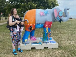 Lucy Anderson with baby Robyn in a sling standing next to an elephant sculpture decorated with trunks and scuba gear