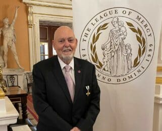 Ray Baglee wearing his award, standing in front of a League of Mercy banner